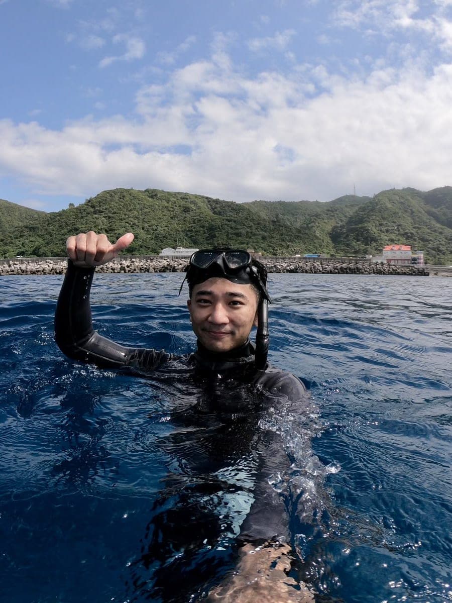 Suyu in a wetsuit and mask at the sea surface, one hand raised, with green hills behind