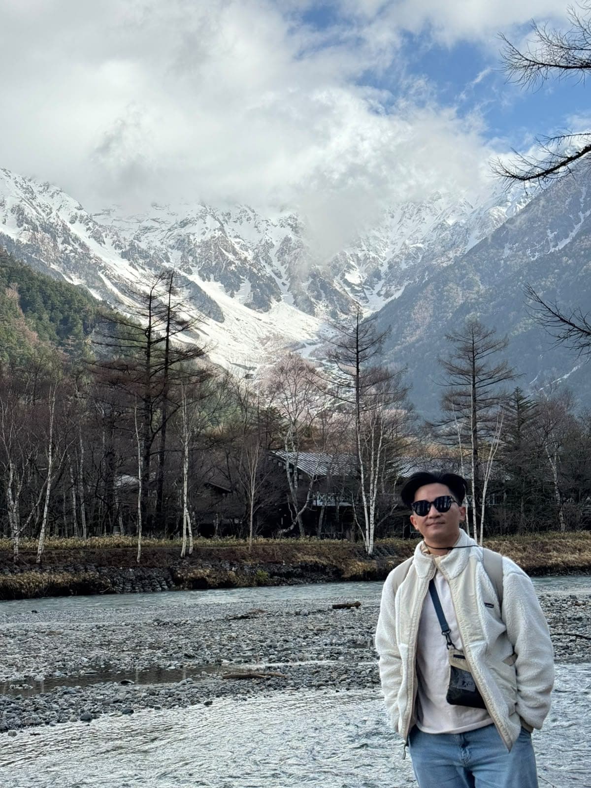 Suyu in sunglasses and a fleece jacket by a rocky river, with snow-covered peaks behind