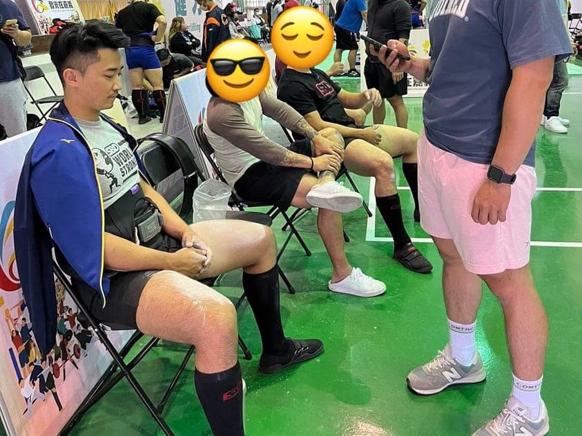 Suyu sitting in the competition warm-up area in a blue jacket, next to other lifters